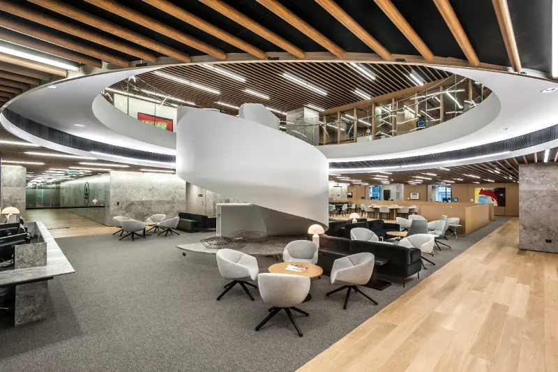 Open-plan workplace lounge beneath a circular timber-batten ceiling with a sculptural white stair