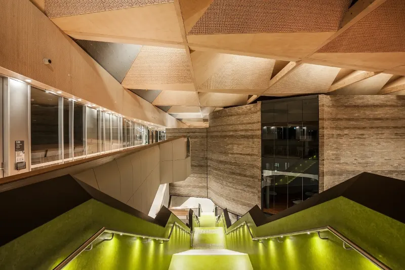 Faceted timber ceiling above a green-lit stair and rammed-earth wall