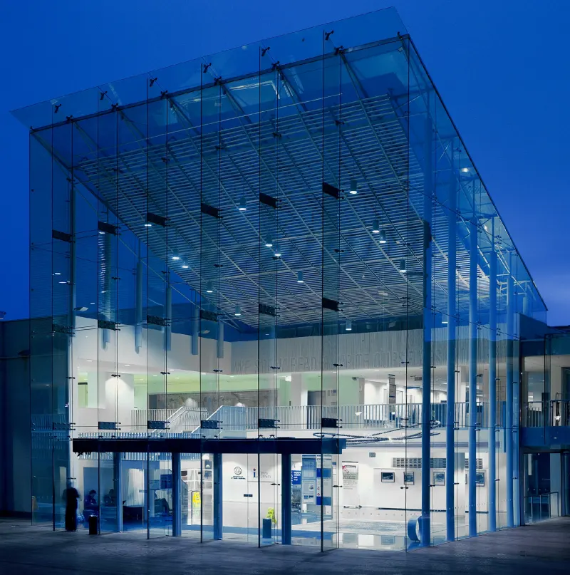 Glazed civic pavilion lit from within at blue hour, structural glass fins to full height